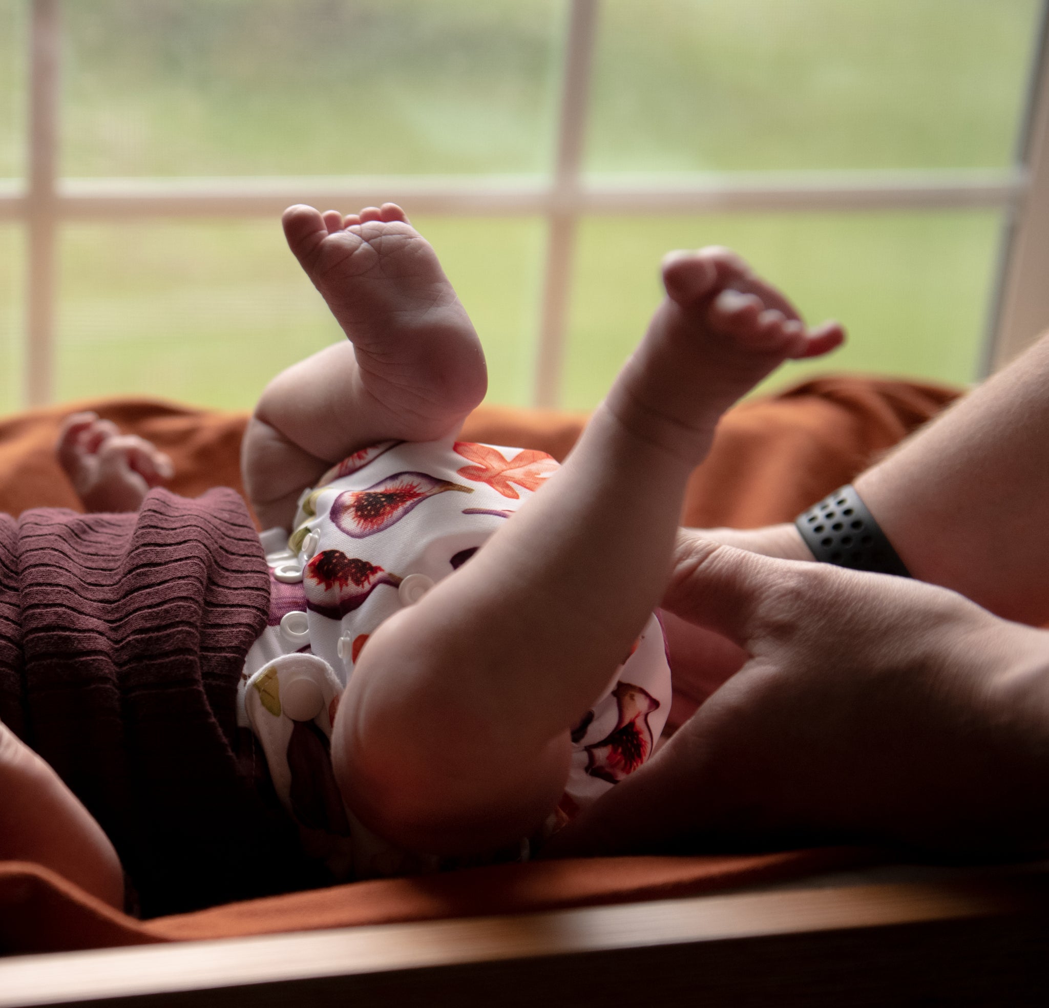-Image of Thirsties Natural Newborn All in One shown in Fig with baby laying on rust colored blanket with window in background