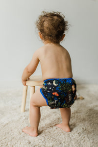 -Image of baby standing on a light colored carpet wearing a Thirsties natural one size pocket diaper shown in Nightlife holding onto a stool. Back of baby is shown.